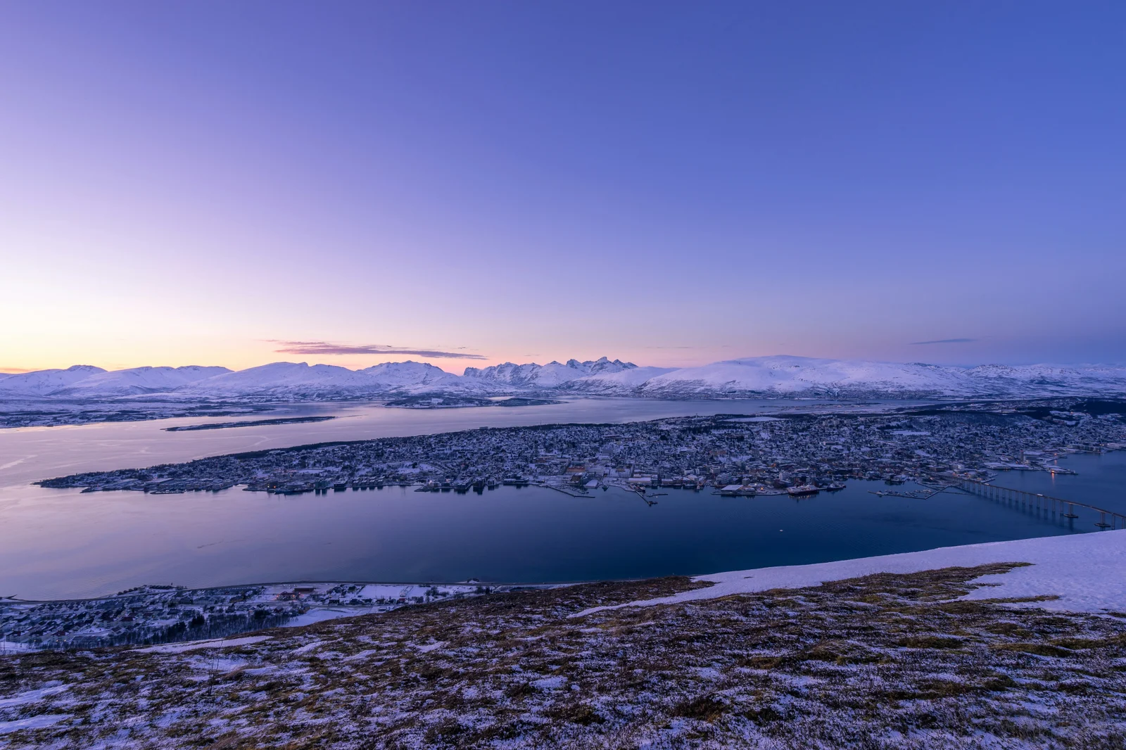 Utsikt over Tromsøya en vinterkveld, med snødekte fjell og Tromsøbrua i blått lys